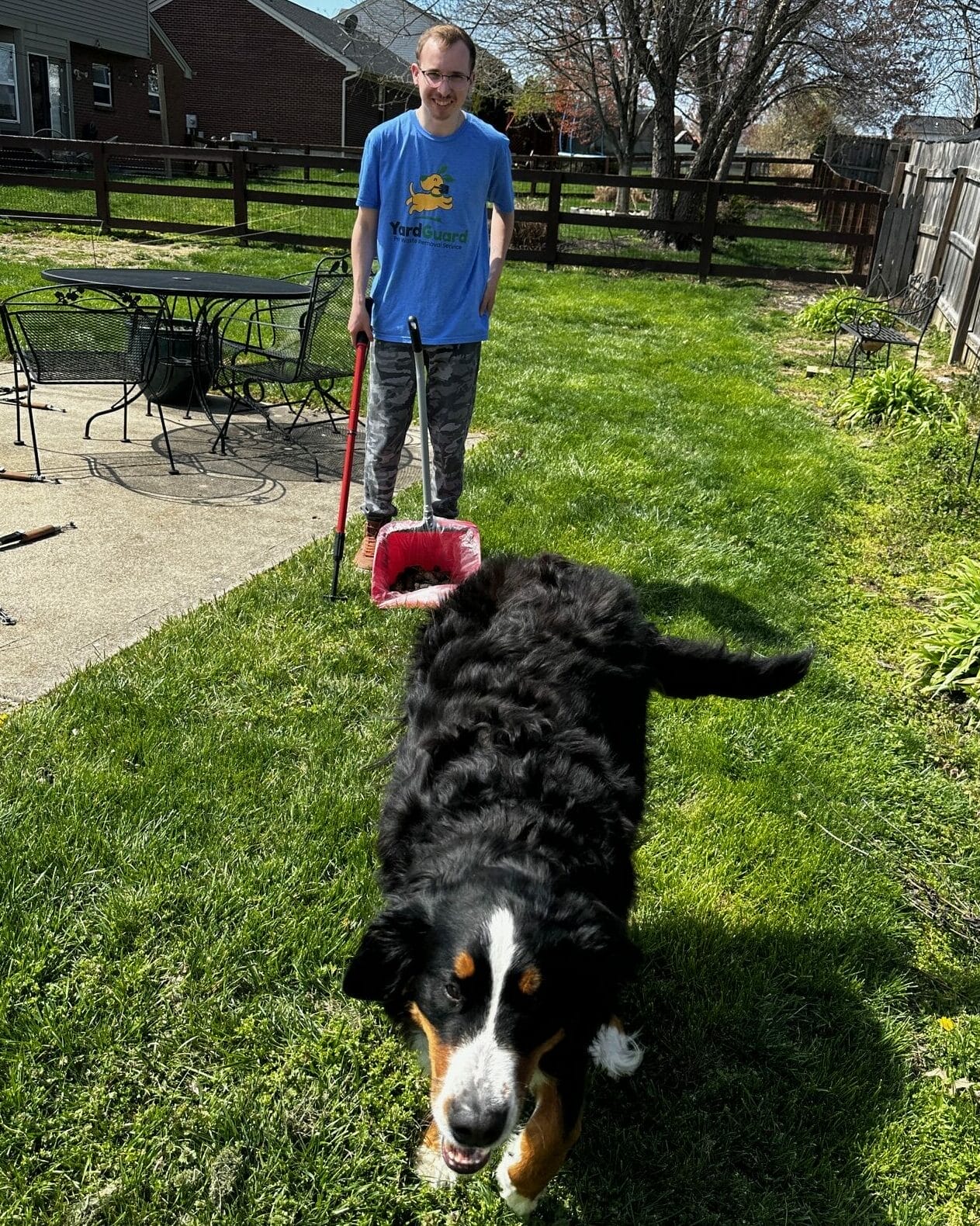 A man in a Yard Guard LLC t-shirt and camouflage pants stands on a grassy lawn holding a red dustpan. A large black and white dog walks towards the camera. The background features a patio with metal furniture and a wooden fence.